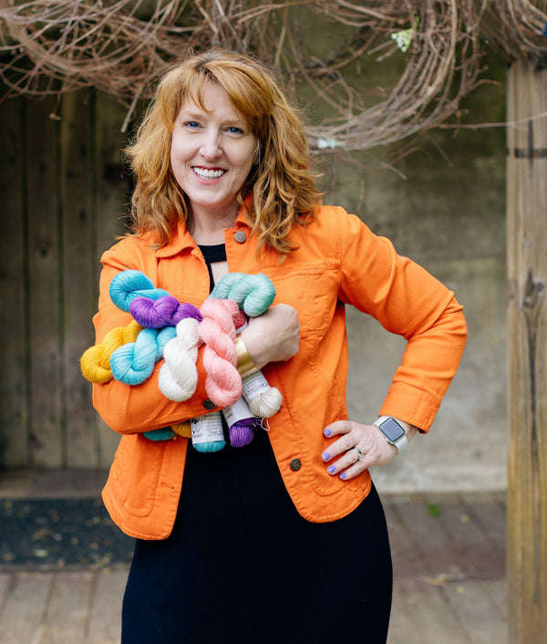 red haired woman wearing orange jacket is smiling with arm full of yarn