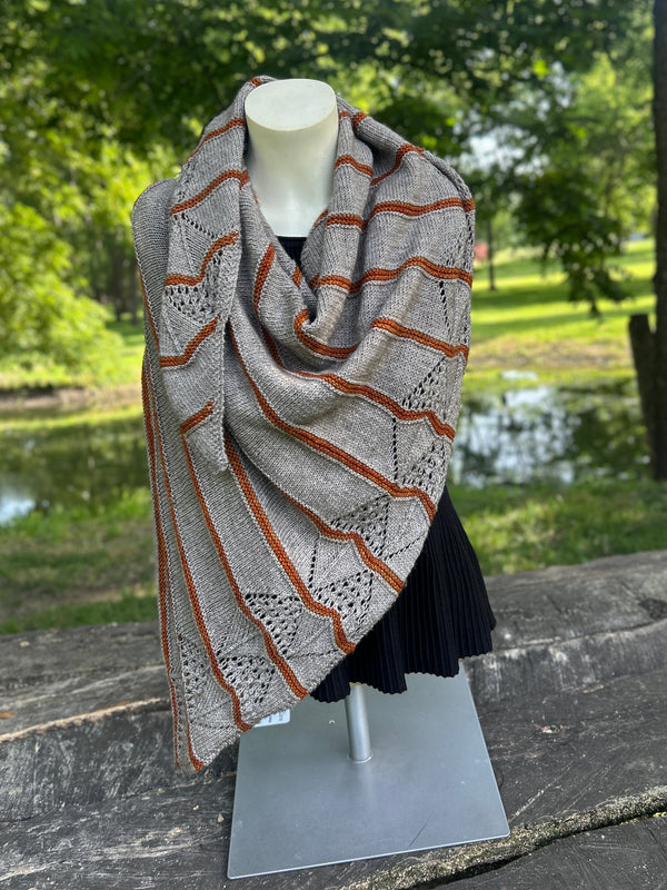 A patterned knit shawl draped on a mannequin torso outdoors on a wooden bench, with a park and pond in the background.